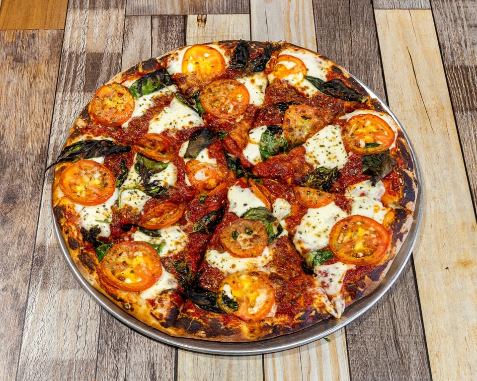 A freshly baked pizza with tomato slices, mozzarella cheese, and basil on a wooden table.