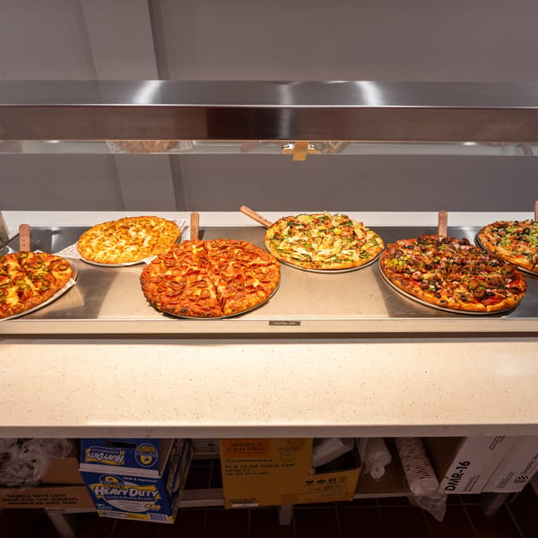 Six different pizzas with various toppings displayed on a heated buffet line.