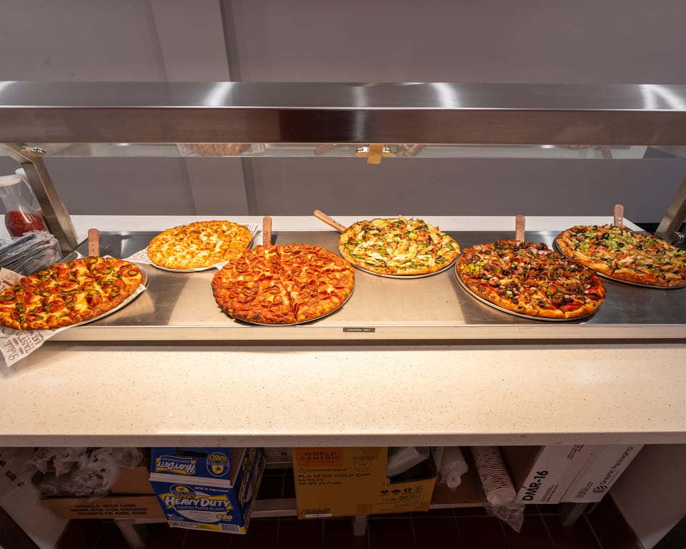 Six different pizzas with various toppings displayed on a heated buffet line.