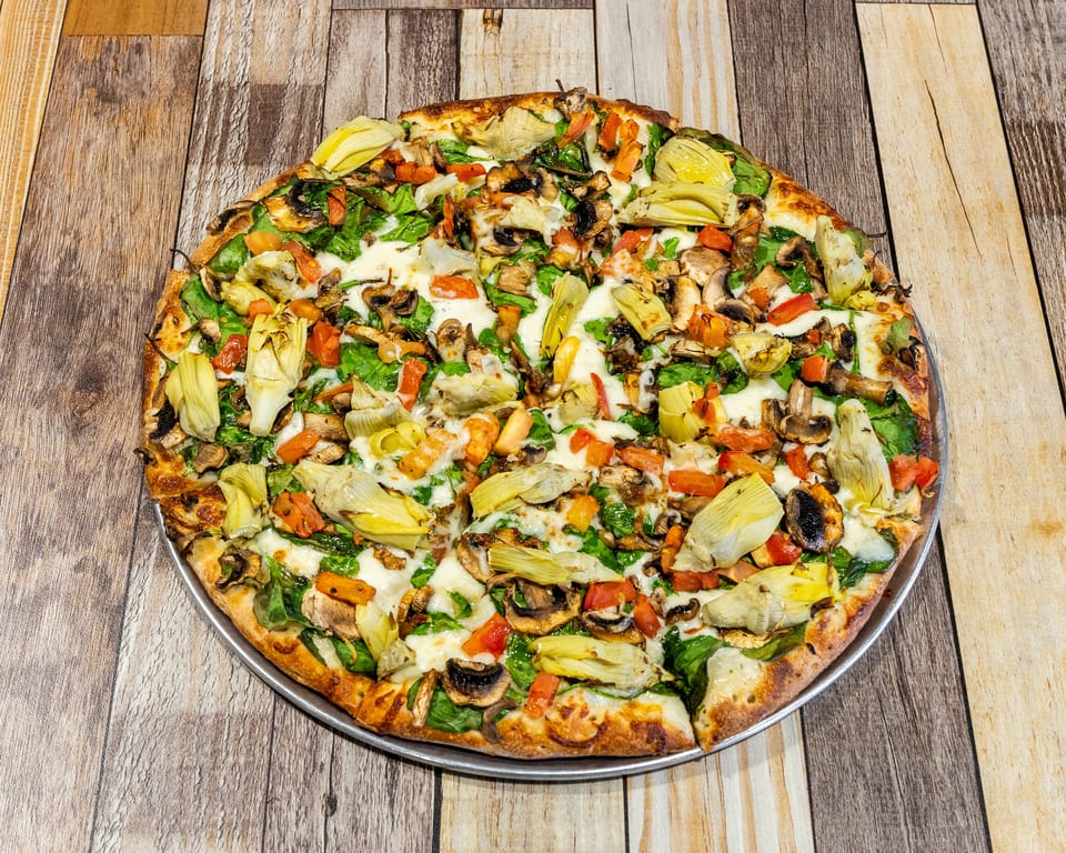 Vegetable pizza with artichokes, mushrooms, spinach, and tomatoes on a wooden table.