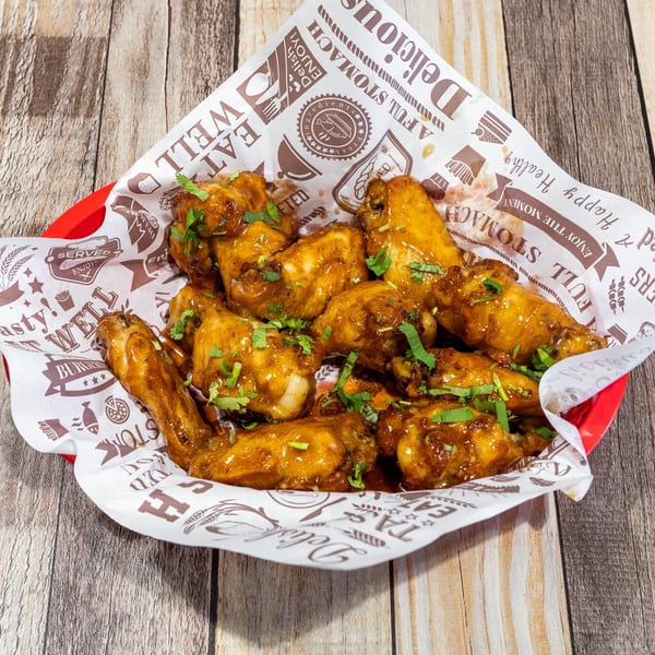 A red basket filled with glossy, golden-brown chicken wings garnished with fresh cilantro, on a wooden table.