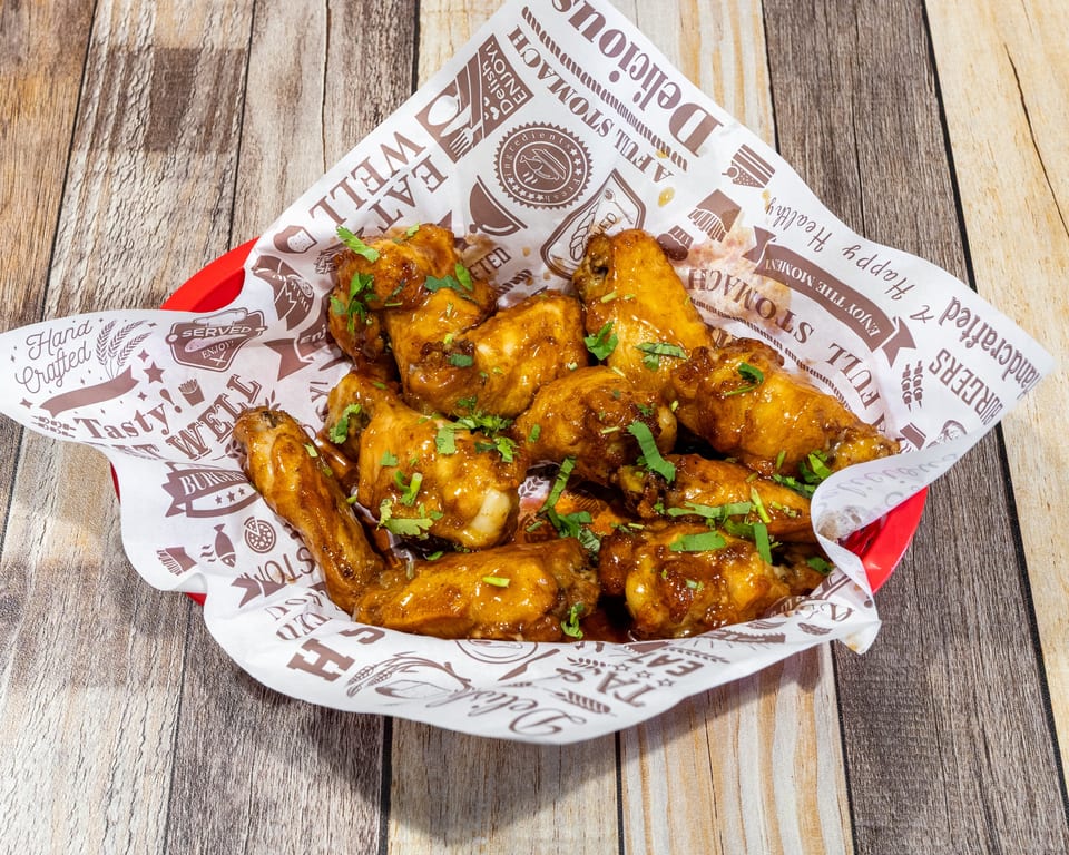 A red basket filled with glossy, golden-brown chicken wings garnished with fresh cilantro, on a wooden table.