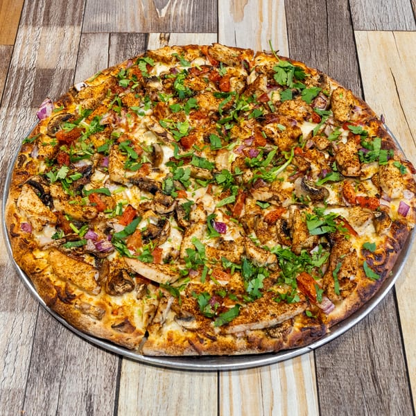 A whole chicken tikka pizza with cilantro, red onions, and mushrooms on a wooden table.