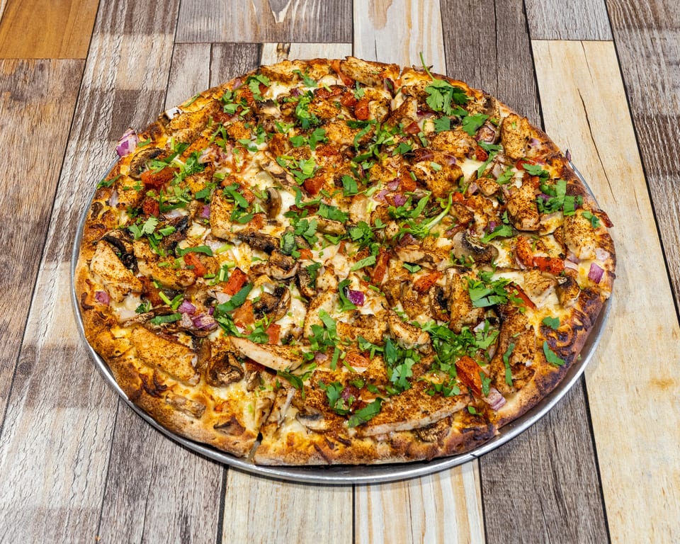 A whole chicken tikka pizza with cilantro, red onions, and mushrooms on a wooden table.