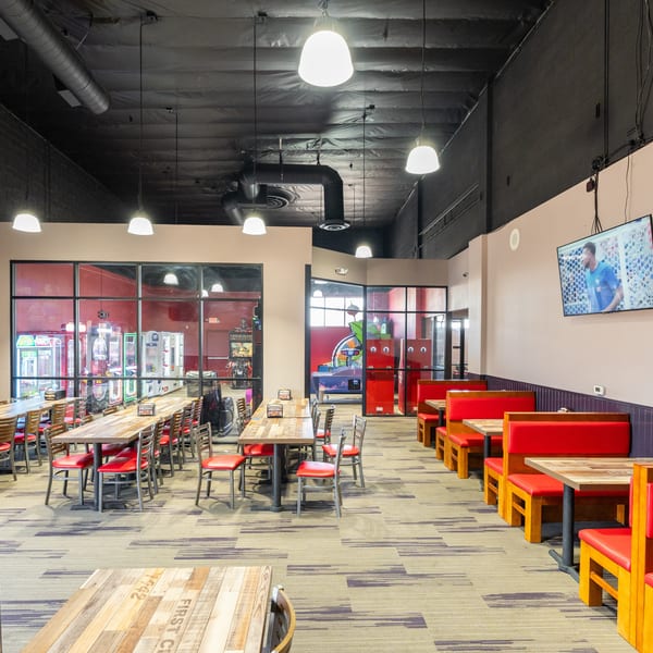 Spacious restaurant dining area with long wooden tables, red chairs, booth seating, and arcade games visible.