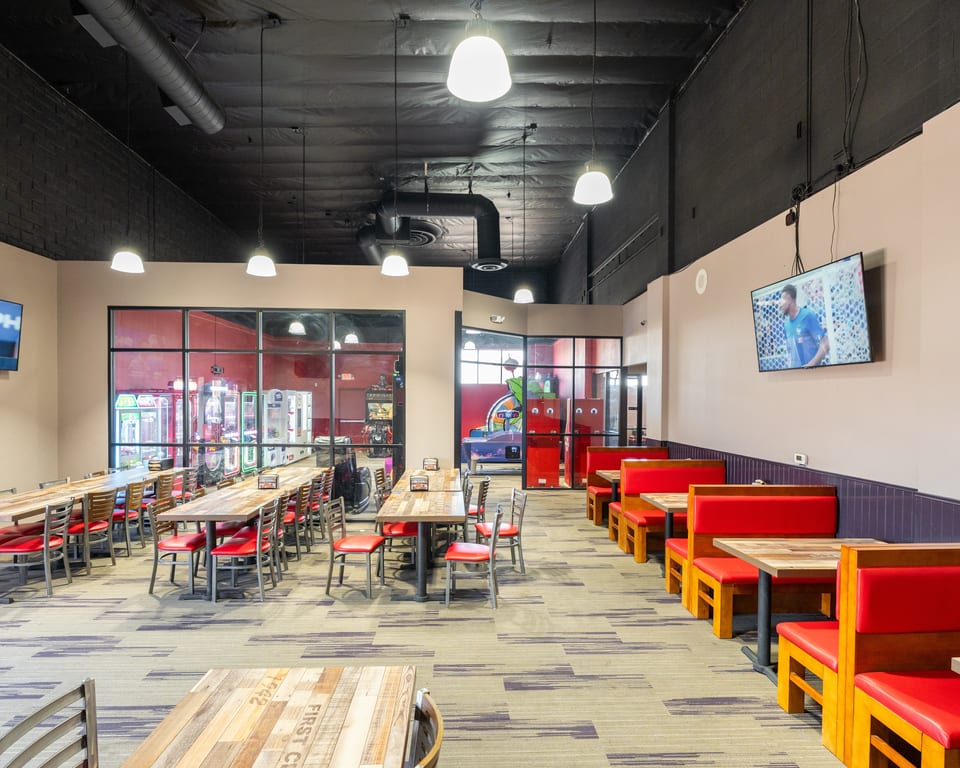 Spacious restaurant dining area with long wooden tables, red chairs, booth seating, and arcade games visible.