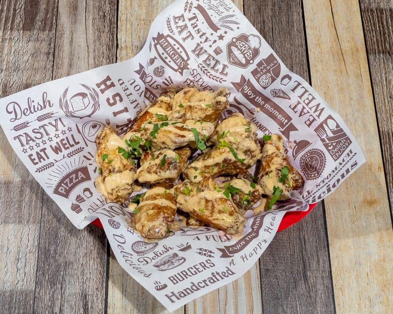 A basket of chicken wings coated in a creamy sauce and garnished with parsley, on a wooden table.