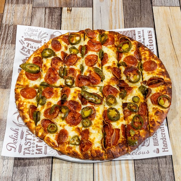 A whole pepperoni and jalapeño pizza, sliced, on branded parchment paper atop a wooden table.