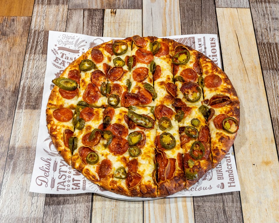 A whole pepperoni and jalapeño pizza, sliced, on branded parchment paper atop a wooden table.