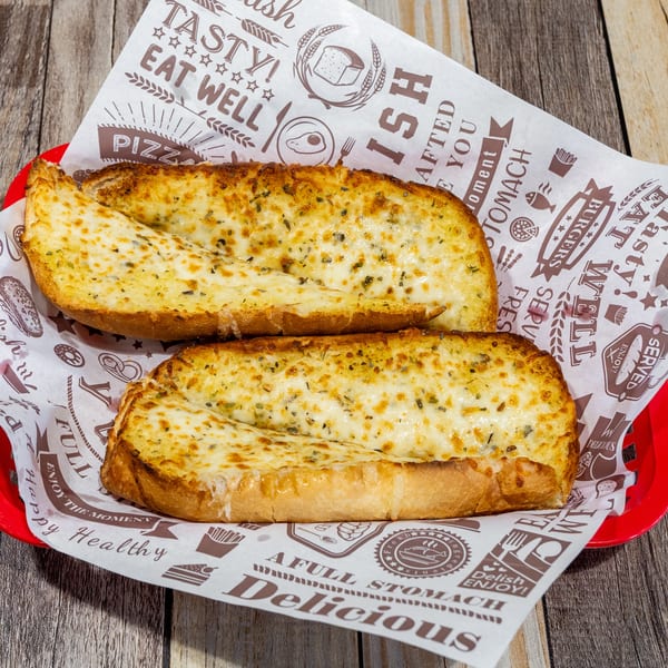 Two slices of cheesy garlic bread with herbs in a red basket lined with patterned paper on a wooden table.