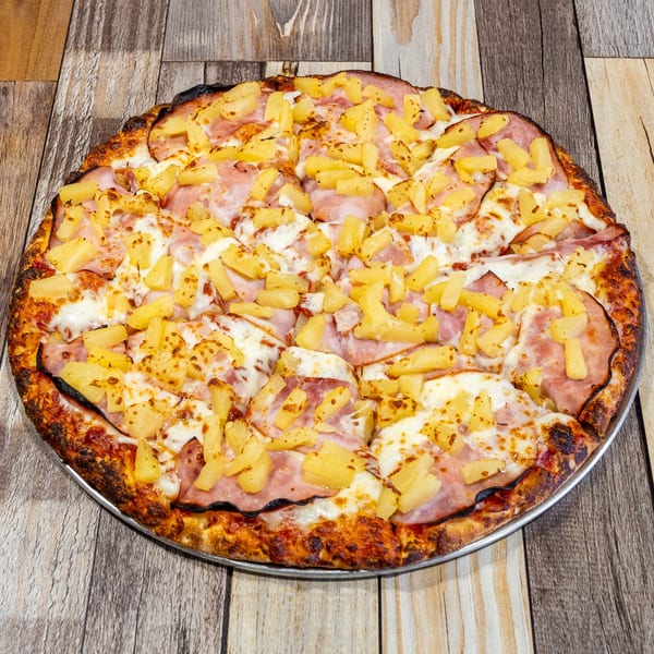 A whole Hawaiian pizza with ham and pineapple on a metal pan, on a wooden table.