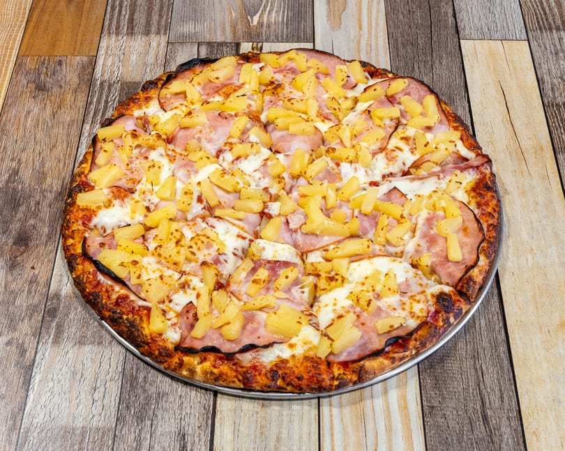 A whole Hawaiian pizza with ham and pineapple on a metal pan, on a wooden table.