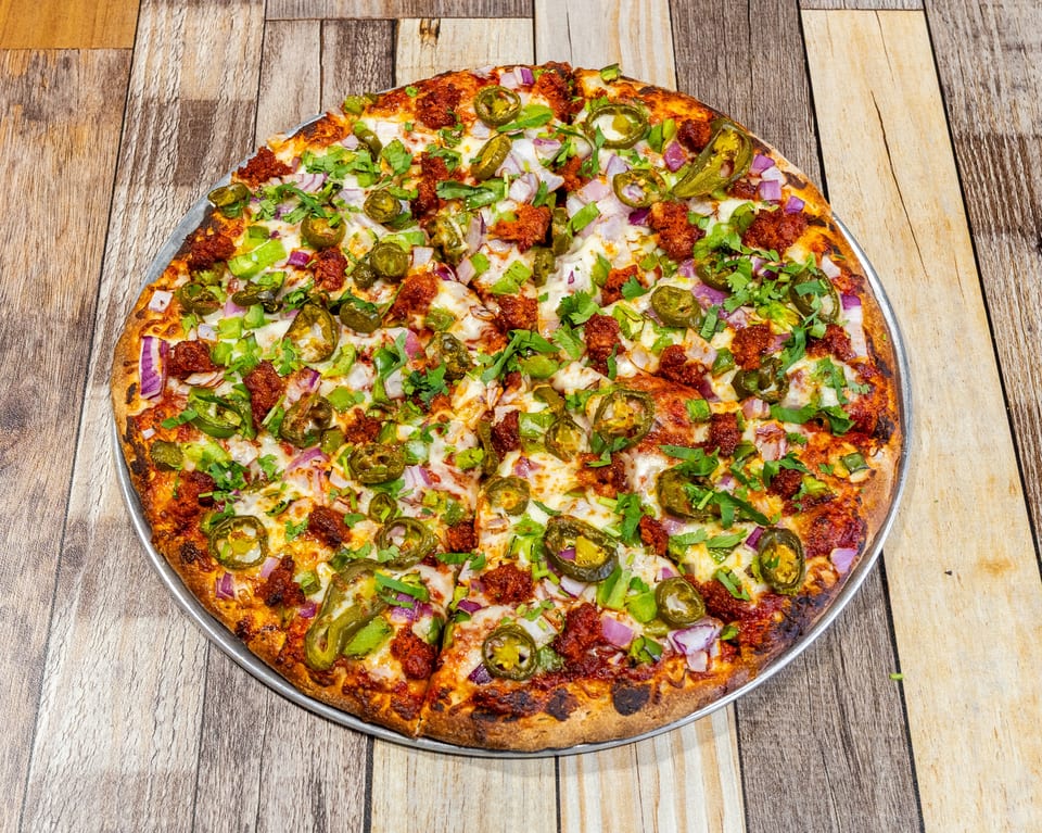 A whole pizza with chorizo, jalapeños, red onions, green peppers, and cilantro on a wooden table.