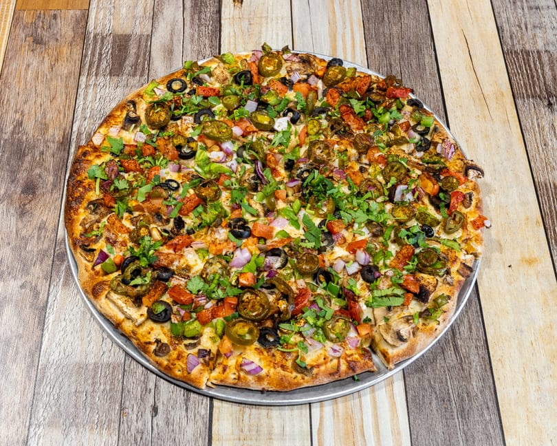 A large, round pizza covered with diverse toppings, including olives, jalapenos, and cilantro.