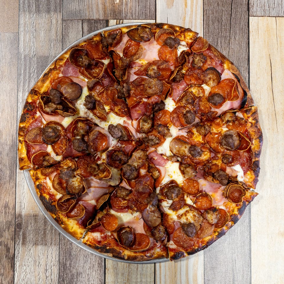 Overhead view of a whole meat lover's pizza with pepperoni, sausage, and ham on a wooden table.