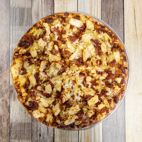 Overhead view of a whole pizza topped with chicken, pineapple, and bacon on a wooden table.