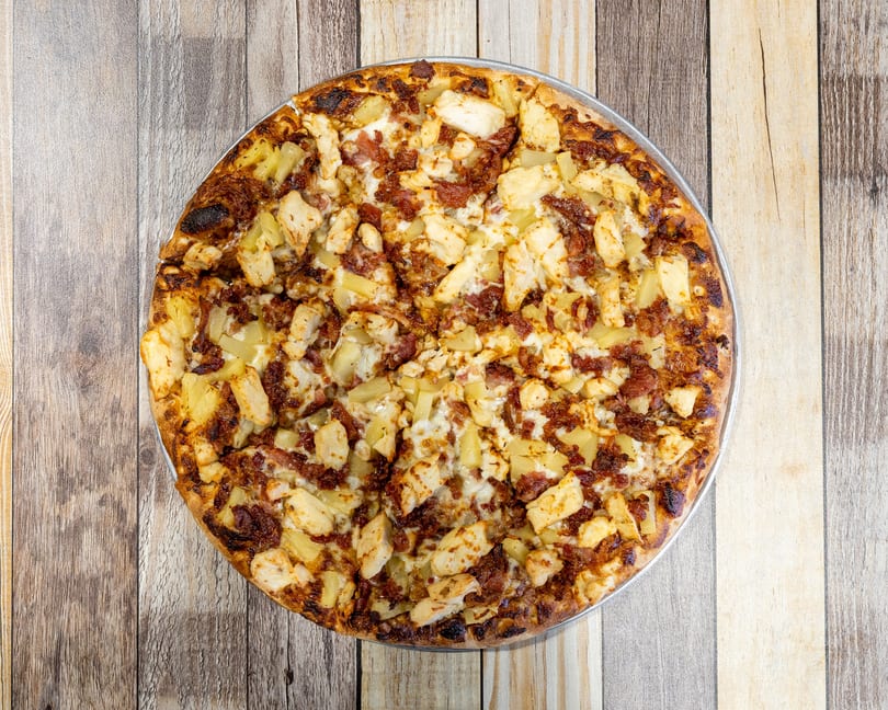 Overhead view of a whole pizza topped with chicken, pineapple, and bacon on a wooden table.