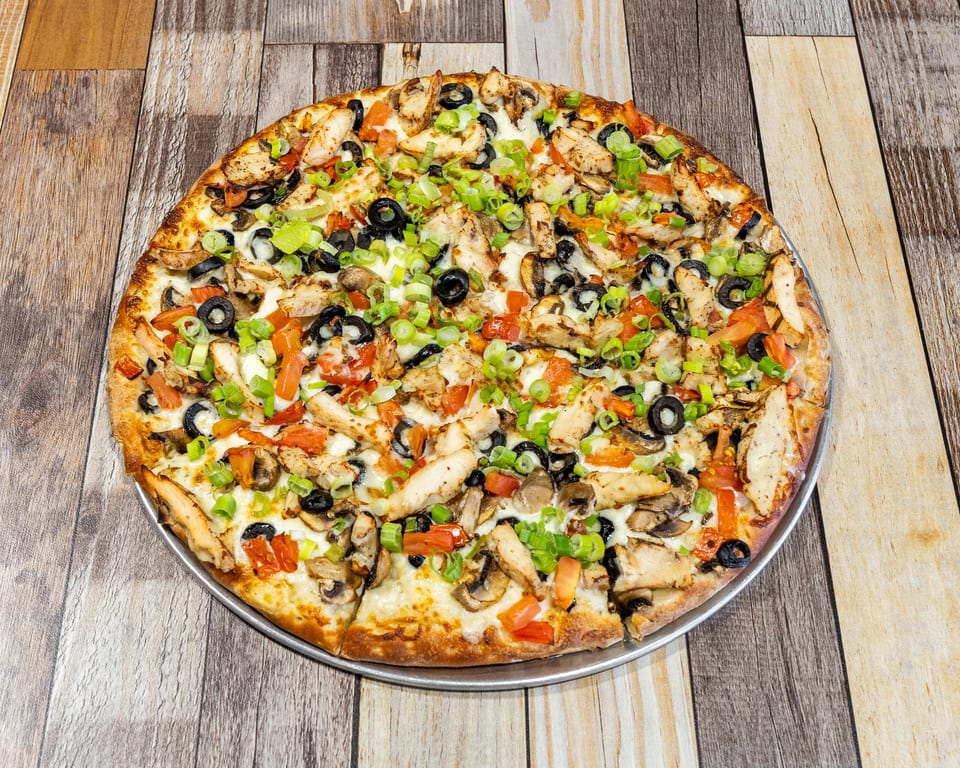 A whole pizza with chicken, black olives, mushrooms, tomatoes, and green onions on a wooden table.