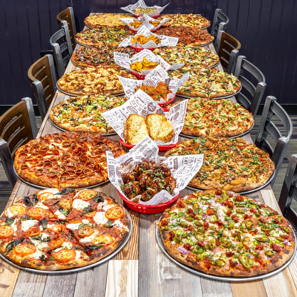 A long table laden with numerous pizzas, garlic bread, fries, and chicken wings in a restaurant setting.