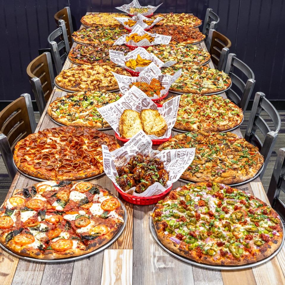 A long table laden with numerous pizzas, garlic bread, fries, and chicken wings in a restaurant setting.