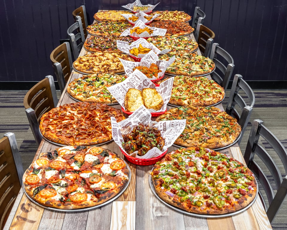 A long table laden with numerous pizzas, garlic bread, fries, and chicken wings in a restaurant setting.