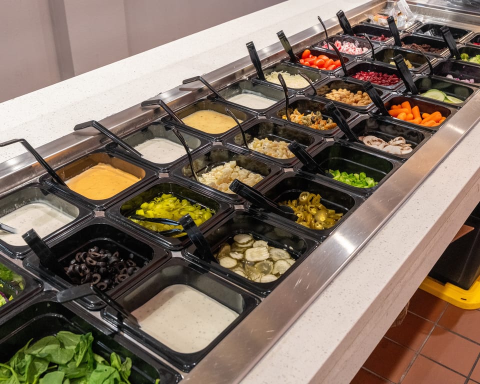 A salad bar with various toppings, dressings, and ingredients in black containers.